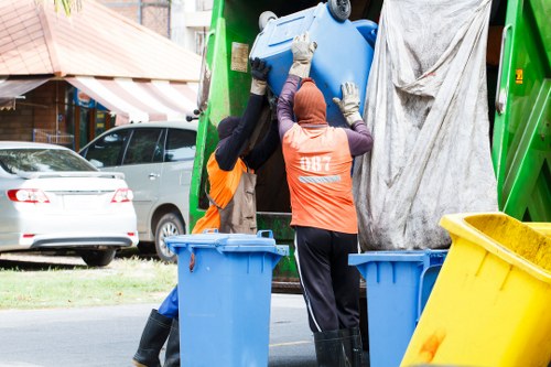 Two waste disposal workers wearing high-visibility orange vests and gloves are collecting a blue plastic wheelie bin from a green waste collection vehicle parked on a street. One worker, wearing black trousers and a pink head covering, is actively lifting the bin, while the other, also in black trousers, is assisting. The waste collection vehicle has a silver body and is positioned at the curb, with a yellow bin and additional blue bins nearby. In the background, there are cars parked along the roadside, residential houses with brick facades, and a small tree. The scene is set during daylight hours, with natural light illuminating the area. This image relates to rubbish removal services in Brockley, London, demonstrating neighborhood waste collection activities facilitated by Commercial Waste Removal Brockley.