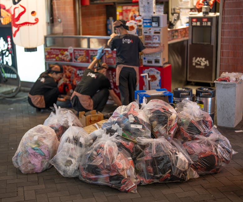A collection of overflowing rubbish bins and scattered waste situated on a paved urban street. The central bin is a large, rectangular, mixed-paper-and-cardo bin made of grey plastic with a blue recyc