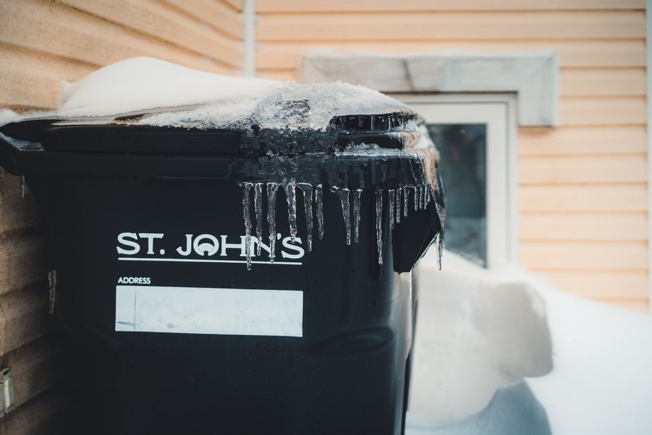 A black outdoor waste bin with the label 'ST. JOHN'S' positioned against a brick wall near the corner of a building during winter. The bin's lid is covered with a thin layer of snow and has ice hangin